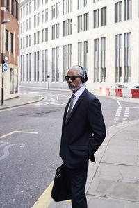 Portrait of elegant senior businessman with sunglasses and headphones standing on sidewalk