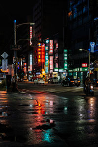 Wet street by illuminated buildings in city during rainy season at night