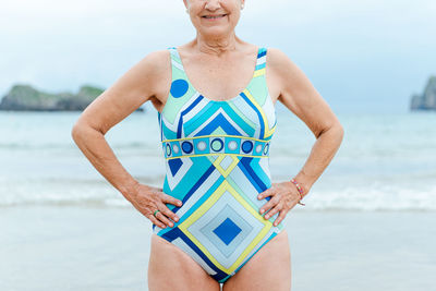 Crop aged female with fit body wearing stylish colorful swimsuit with geometric print standing with hands on waist against sea in summer day