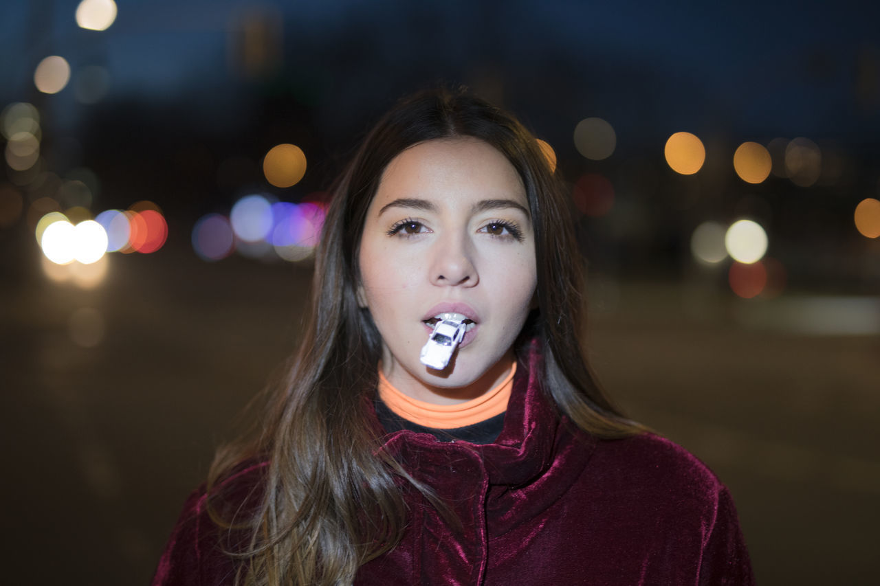 PORTRAIT OF BEAUTIFUL YOUNG WOMAN STANDING IN ILLUMINATED CITY