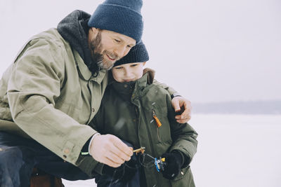 Smiling mature man in warm clothing showing fishing rod to son