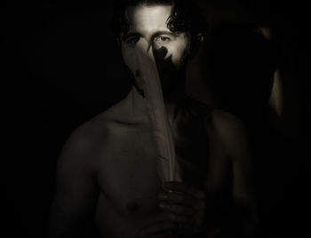 Portrait of shirtless young man holding feather while standing against black background