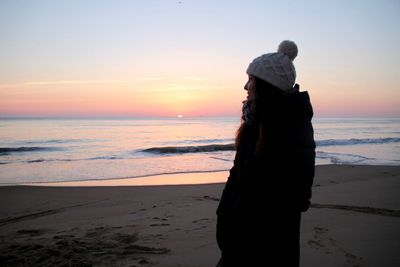 Side view of woman in warm clothing at beach against sky during sunrise