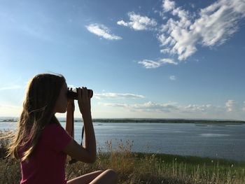Woman looking at sea through binoculars against blue sky