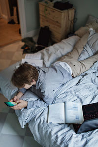 High angle view of social media addicted teenage boy using mobile phone while lying by books in bedroom