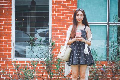 Portrait of woman holding drink standing against brick wall