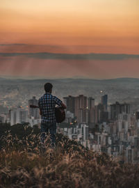 Rear view of man playing guitar while standing against cityscape and sky during sunset