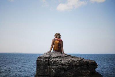 Rear view of female hiker with backpack sitting on rock against sea and sky at bruce peninsula national park