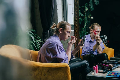 Man applying make-up sitting on chair by mirror