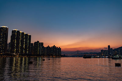 Illuminated city by river against sky during sunset