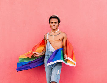 Portrait of gay man holding rainbow flag against pink wall
