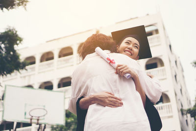 Cheerful woman in graduation gown embracing mother while standing outdoors