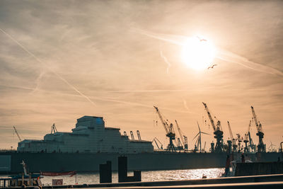 Silhouette cranes at harbor against sky during sunset