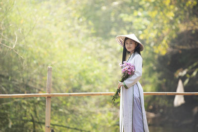 Portrait of woman holding flowers while standing by railing against trees
