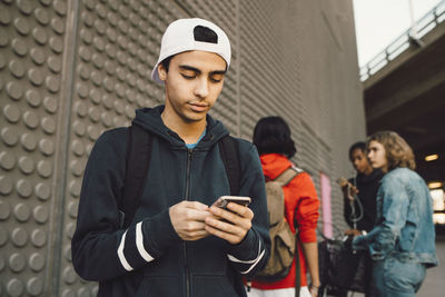 Young man using mobile phone with friends standing in background on footpath