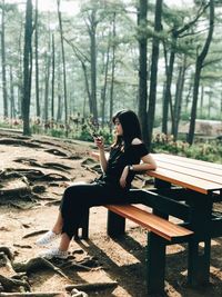 Full length of young woman using smart phone while sitting on picnic table in forest