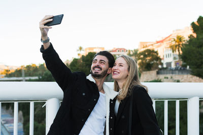 Joyful young multiracial couple in casual wear taking selfie on mobile phone while standing together on bridge with green trees and city buildings in background