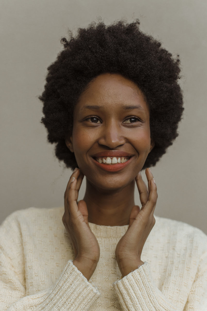 portrait, hairstyle, afro, smiling, human hair, one person, curly hair, looking at camera, happiness, adult, women, headshot, studio shot, indoors, front view, emotion, cheerful, gray, teeth, human face, smile, gray background, young adult, person, black hair, female, human head, nose, clothing, enjoyment, long hair, positive emotion, casual clothing, short hair, brown, close-up, individuality, colored background, skin