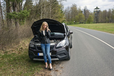 Full length of fashionable young woman standing by breakdown car on roadside