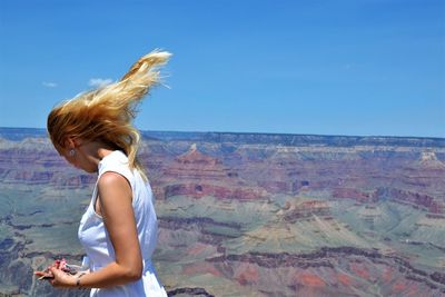 Side view of woman with tousled blond hair standing at grand canyon national park against blue sky