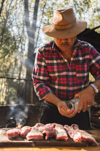 Adult argentinian male with mustache wearing beige hunter hat and checkered shirt salting meat on cutting board while preparing grill in picnic in nature