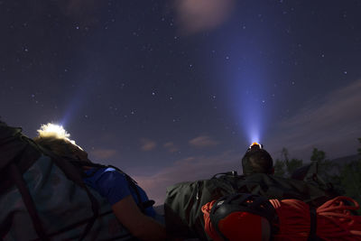 Two people laying down on backpack under the stars with headlamps