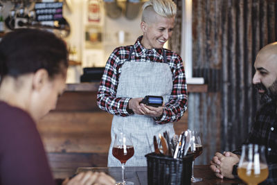 Smiling female bartender holding credit card reader while talking to customers at table in bar