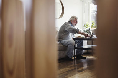 Senior man using digital tablet while having breakfast seen through wooden columns