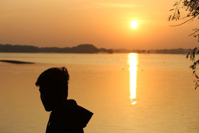 Silhouette boy standing by sea against sky during sunset