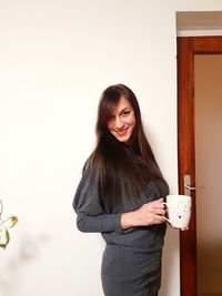 Portrait of smiling woman holding coffee cup standing against wall at home