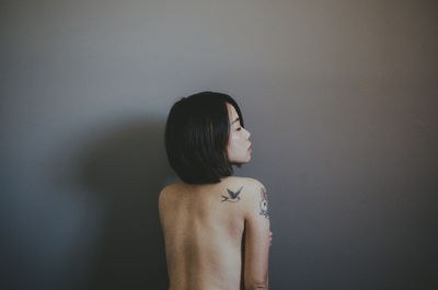 Rear view of shirtless woman standing against grey wall