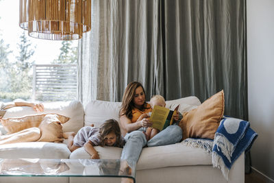 Mother reading book sitting with toddler son by daughter on sofa at home