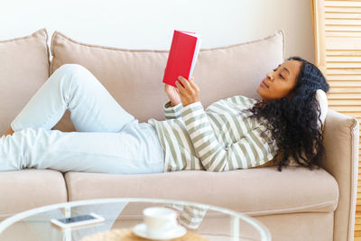 African-american lying down on sofa and reading book in red cover. slow paced lifestyle