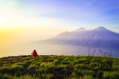 Rear view of man sitting on mountain against sky during sunset