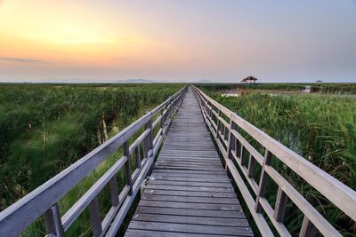 Wooden boardwalk amidst field against sky during sunset