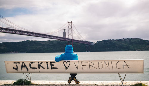 Woman sitting on bench while looking at bridge over river against sky