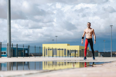 Shirtless man standing by puddle on street against cloudy sky