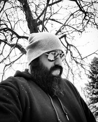 Bearded man wearing knit hat and sunglasses against bare tree