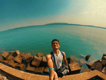 Portrait of young man sitting by sea against sky