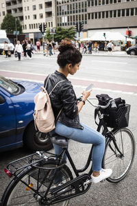 Full length of woman with bicycle using mobile phone while standing at city street