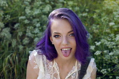 Portrait informal teenage girl with purple hair and piercing shows tongue on green field in summer