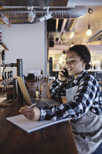 Smiling female manager talking on mobile phone while using laptop at bar counter