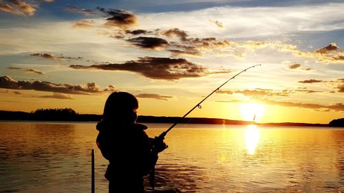 Rear view of silhouette girl fishing in lake against sunset sky