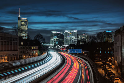 Light trails on road amidst buildings against sky at night