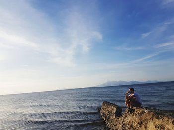 Man sitting on rock looking at sea against sky