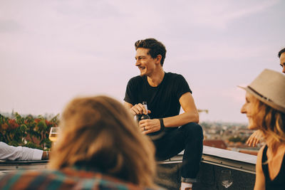 Smiling man looking away while enjoying beer with friends at rooftop party against sky