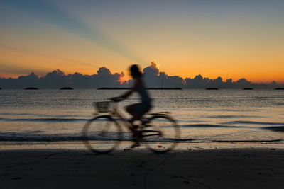 Silhouette person riding bicycle on beach against sky during sunset