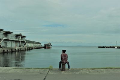 Rear view of teenage boy looking at sea while sitting on bollard against cloudy sky
