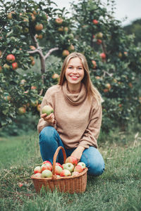 Portrait of smiling young woman holding apple while crouching by basket against trees