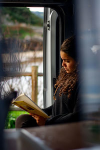 Woman reading book while sitting by window at home